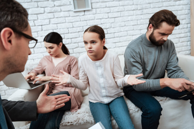 Complex Family Law Dispute Mediation - Girl tries to communicate with a family member while sitting on a sofa. Dad and Mom do not want to talk to each other.